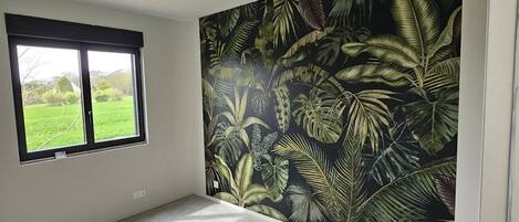 Ett rum med ett stort fönster, en vägg med ett tropiskt bladmotiv och ett klinkergolv.