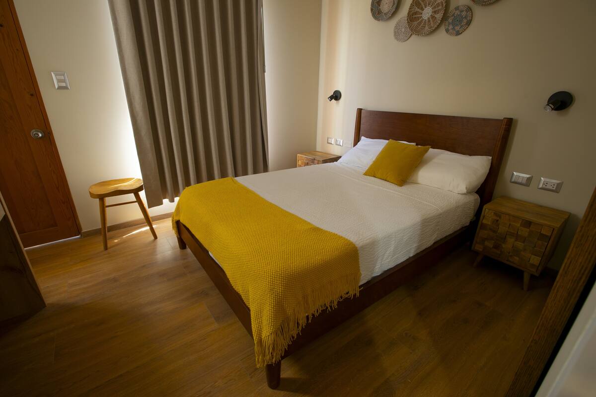 A bedroom with a wooden bed, a yellow throw blanket, a wooden nightstand, and a small stool.