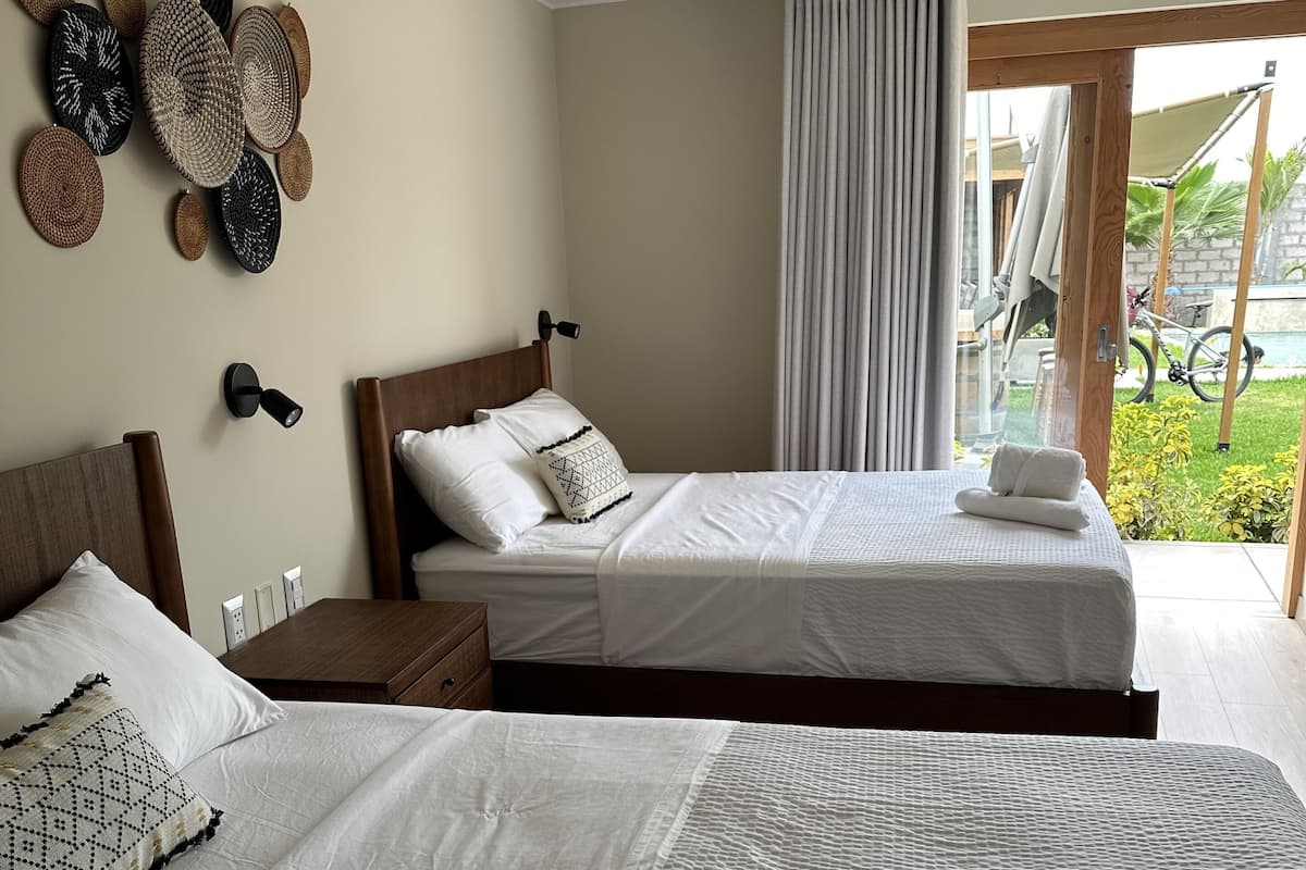 A hotel room with two beds, a wooden headboard, a wall-mounted lamp, and a view of a garden through a sliding door.