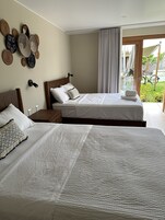 A hotel room with two beds, a wooden headboard, a wall-mounted lamp, and a view of a garden through a sliding door.