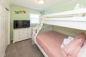 Une chambre d’enfant avec des lits superposés, une commode, un téléviseur et une fenêtre avec des stores.