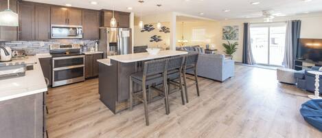 Una cocina moderna con una isla central, un comedor con un taburete de bar y una sala de estar con sofá y televisor.