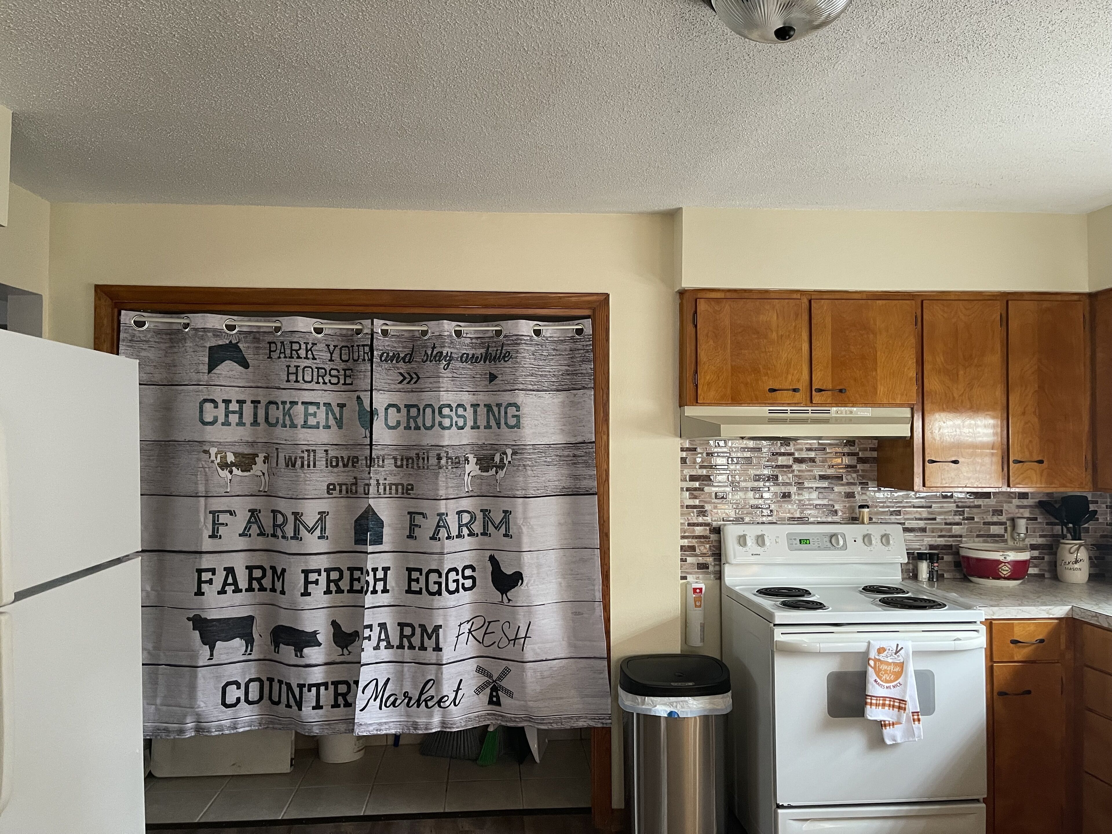 Une cuisine avec des meubles de rangement en bois, un fourneau blanc et un rideau orné d’un motif ou d’un texte sur le thème de la ferme.
