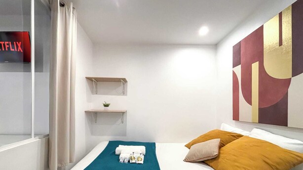 Ein modernes Hotelzimmer mit Bett, Kissen in Türkis und Senfgelb sowie einer türkisfarbenen Tagesdecke und einem an der Wand befestigten Regal mit Pflanze.