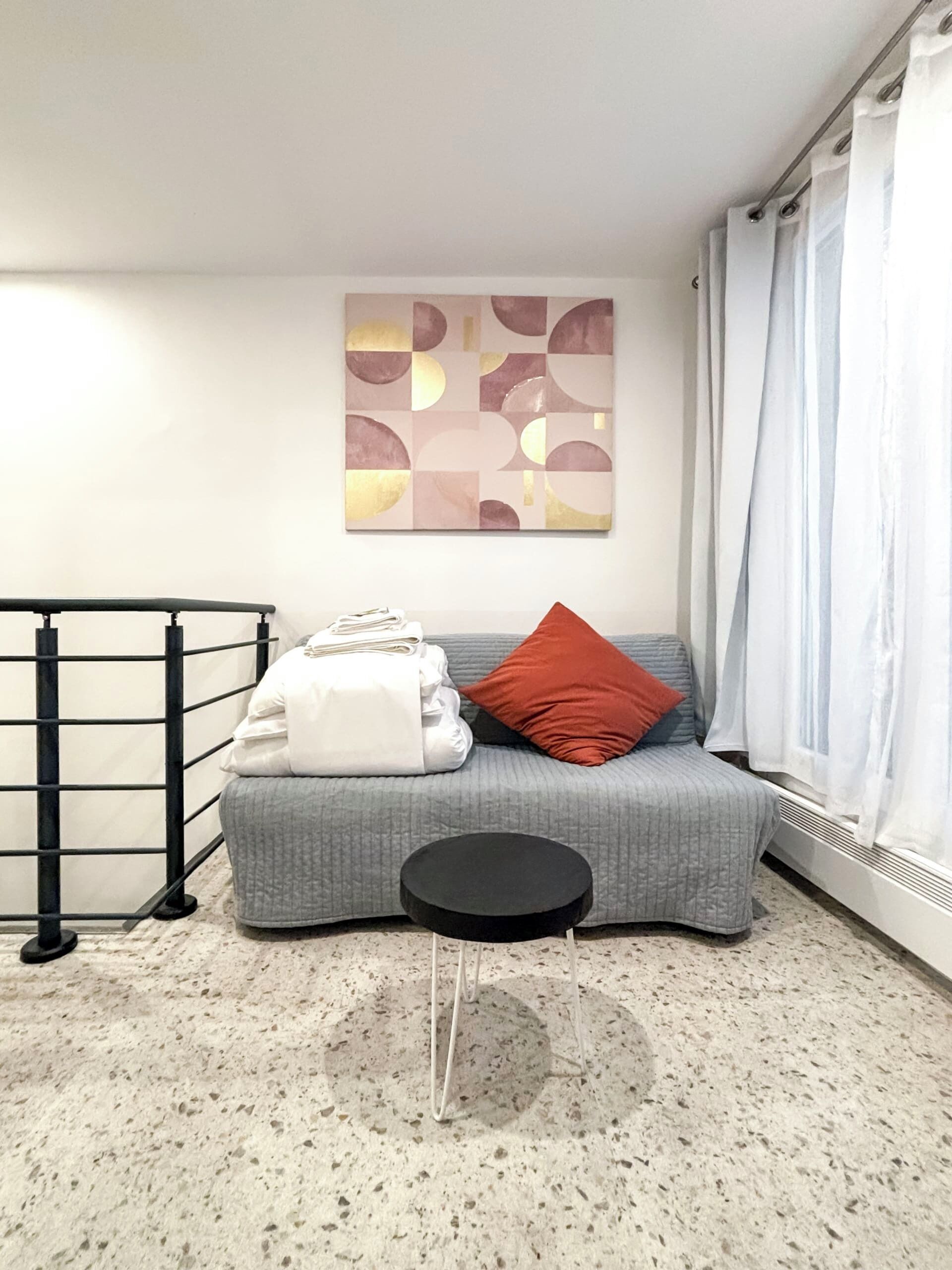 Un salon moderne avec un canapé gris, un coussin rouge, une couverture blanche et une table d’appoint noire. Un grand tableau abstrait est accroché au mur, et une fenêtre avec des rideaux blancs est visible.