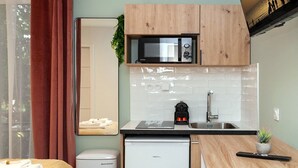 Eine kompakte Küchenzeile mit Mikrowelle, Spüle und kleinem Kühlschrank. Ein Holzbett und ein kleiner Tisch mit Hocker sind ebenfalls vorhanden.