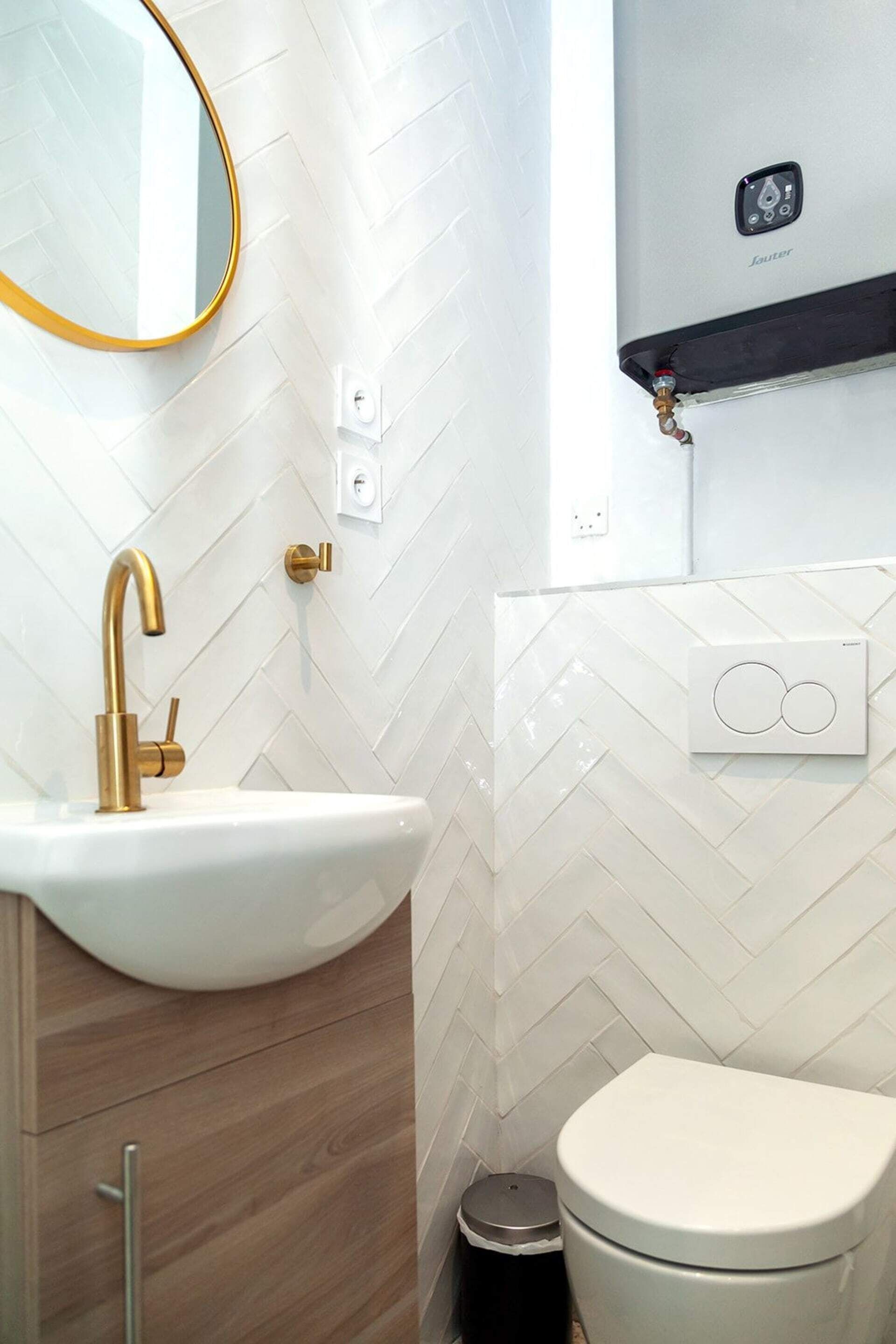 A bathroom with a white herringbone tile backsplash, a gold faucet, a white sink, a toilet, and a mirror with a gold frame.