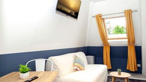 Habitación de hotel con cama, una mesita con un control remoto, una planta en maceta, una ventana con cortinas y un televisor montado en la pared.