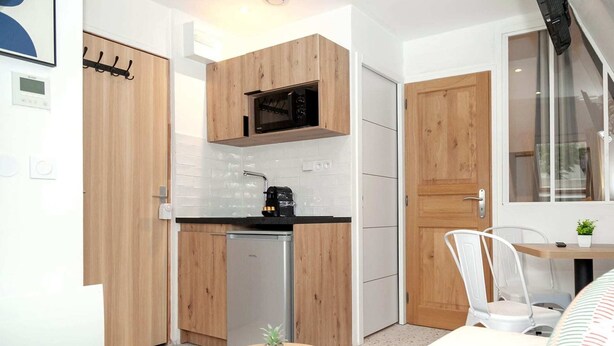 Cocina pequeña con armarios de madera, microondas y lavavajillas. Hay una mesa redonda con una planta y una silla blanca.