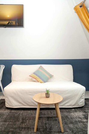 Un sof á blanco con un cojín colorido, una mesita de madera con una planta, y un cuadro de una puesta de sol en la pared.