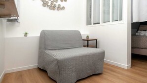 A corner room with a grey upholstered chair, a small table with a plant, and a window with blinds. - David D Angers 29 - One Bedroom Apartment, Sleeps 3 (Paris)