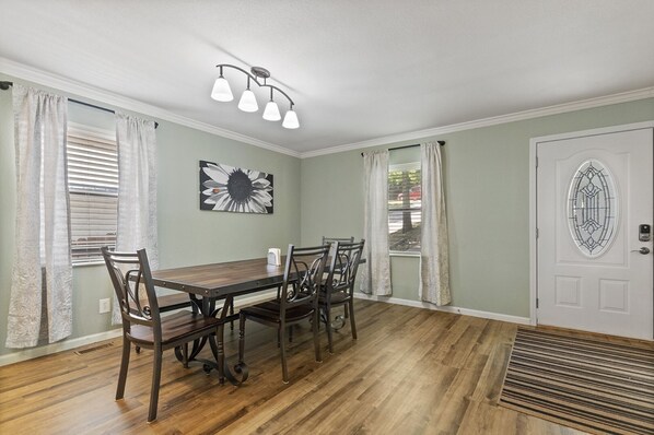 Un coin repas avec une table et des chaises en bois, un luminaire au plafond, une porte avec un panneau de verre et une fenêtre avec des stores.