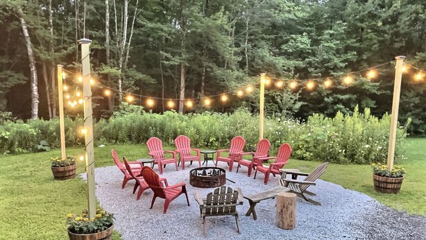 An outdoor seating area with red chairs and a fire pit, surrounded by a gravel path and string lights.