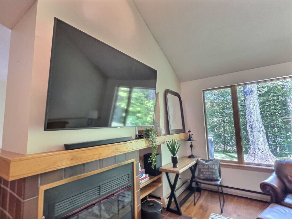 A living room with a fireplace, a television mounted above it, a window with a view of trees, and a small table with a lamp.