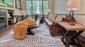 A cozy living room with a fireplace, a sofa, a coffee table, and a TV mounted on the wall. - SugarBear- Pool, AC, Sauna, Gym, Tennis! (Killington)