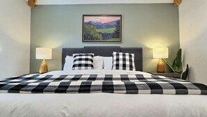 Un lit bien fait, recouvert d’une couvre-lit à carreaux, deux lampes de chevet, un tableau représentant un paysage encadré au-dessus du lit et une plante en pot sur la table de chevet.