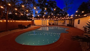 A swimming pool with string lights, a covered seating area, and a building with a door and windows.