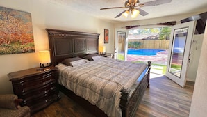 A bedroom with a large bed, wooden headboard, a ceiling fan, a nightstand, a lamp, a dresser, and a painting on the wall.