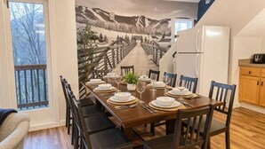 A dining room with a long table set for eight, wooden chairs, a refrigerator, and a kitchen area with wooden cabinets. - BearQuad- AC, Pool, Tennis, Close to Resort/Biking (Killington)
