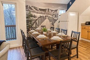 Comedor con una larga mesa dispuesta para ocho personas, sillas de madera, un refrigerador y una zona de cocina con armarios de madera.
