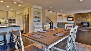 A modern kitchen with a central island, stainless steel appliances, and a dining area with a wooden table and white chairs. - Beartopia- Hot Tub, Views, 2 Primary Suites! (Killington)