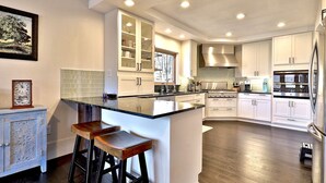 A modern kitchen with white cabinetry, a large island with stools, stainless steel appliances, and recessed ceiling lights. - Beartopia- Hot Tub, Views, 2 Primary Suites! (Killington)