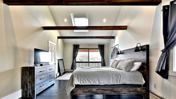 A bedroom with a large bed, a dresser, a TV, and a window with curtains. - Beartopia- Hot Tub, Views, 2 Primary Suites! (Killington)