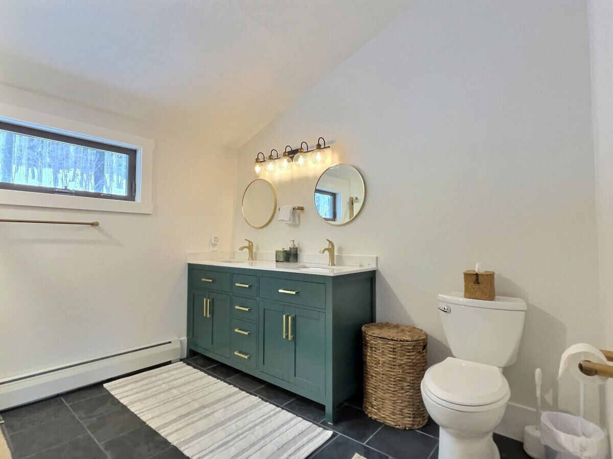 Een badkamer met een blauwgroene wastafel, twee ronde spiegels, een wit toilet en een klein rieten mandje.