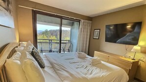 A hotel room with a large bed, a bedside table with a lamp, a TV mounted on the wall, and a balcony with a view of trees. - Rockstar - Pool, Tennis, Fireplace, Pickleball (Killington)