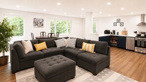 A modern living room with a sectional sofa, ottoman, and a kitchen area with appliances. - CubsCorner - Pool, Sauna, Gym, Close to Mountain! (Killington)
