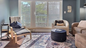 A living room with a large sliding glass door, a sofa, a chair, a small table, and a rug. - Moose Tracks Lodge - Pool, Hot Tub, Golf, Sauna (Killington)