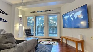 Uma sala de estar com um sofá cinzento, um banco de madeira, uma TV fixada na parede e uma janela grande com vista para o exterior.