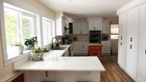 A modern kitchen with white cabinetry, a central island with stools, and a window with plants. - Large Kitchen, Hot tub, Game & Arcade Room (Hayden)