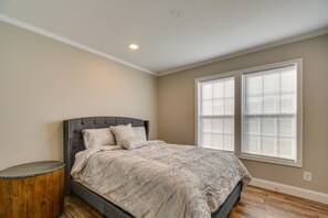 A bedroom with a bed, a round wooden nightstand, and two windows with blinds.
