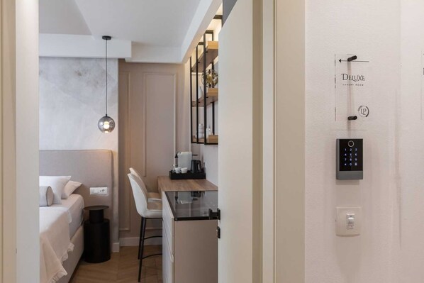 A modern hotel room with a bed, a desk, and a chair. There is a wall-mounted digital lock and a small trash bin.