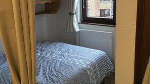 Une petite chambre avec un lit simple, une couvre-lit à motifs, une fenêtre avec des rideaux et une vue sur un bâtiment à l’extérieur.