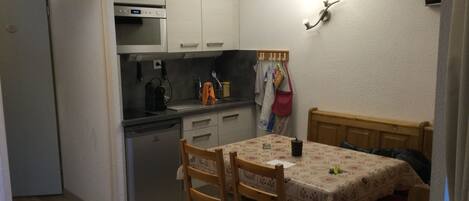 Eine kompakte Küche mit Kühlschrank, Backofen und Geschirrspüler. Ein Esstisch mit Stühlen und ein kleines Bett mit roter Decke sind vorhanden.