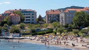 Ein Strand mit Personen, die sonnenbaden und schwimmen, umgeben von Gebäuden und Grünflächen.
