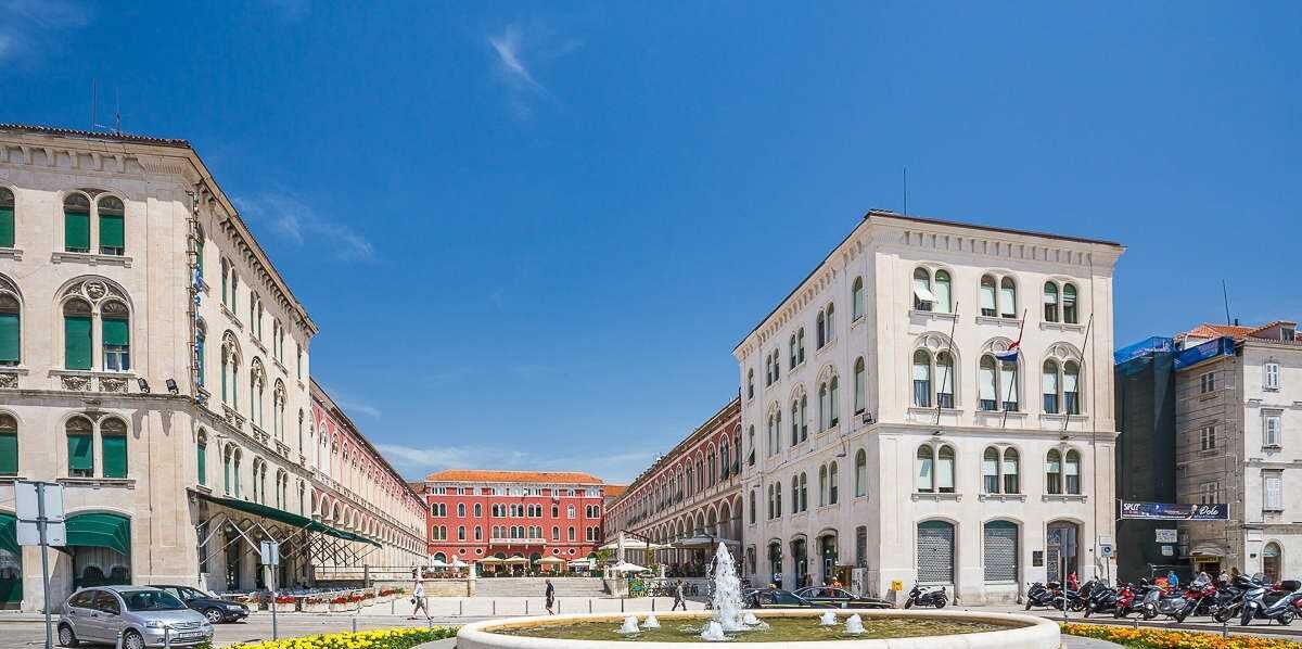 Uma praça histórica com arquitetura clássica, uma fonte e um edifício vermelho ao fundo.