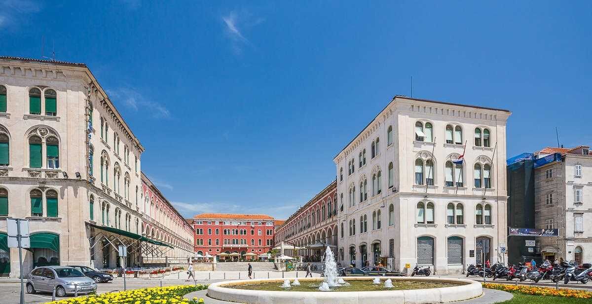 A historic square with classical architecture, a fountain, and a red building in the background.