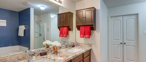 Une salle de bain avec un double lavabo, un grand miroir et une douche. On y trouve des armoires en bois et une serviette rouge accrochée au mur.