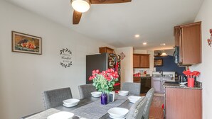 Un comedor con una mesa larga dispuesta para seis personas, una cocina al fondo y una pared decorativa con una pintura de frutas y un letrero que dice 'Grateful Blessed'.