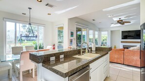 A modern kitchen with granite countertops, a double sink, and a built-in oven. There is a dining area with a glass table and chairs, and a large window offering a view of the outdoors. - Cathedral City Home w/ Private Pool - Near Golf (Cathedral City)