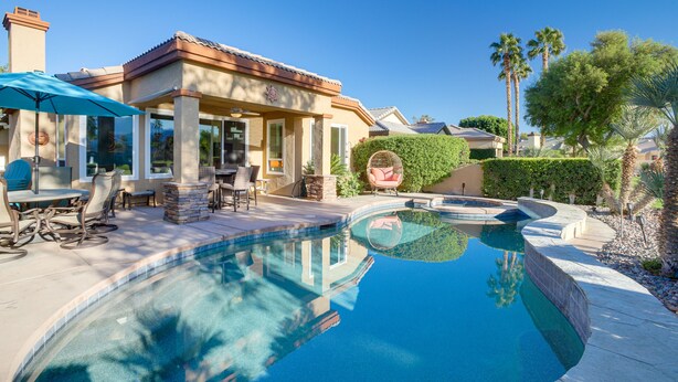 A swimming pool with a curved edge, a covered patio area with outdoor furniture, and a well-maintained garden with palm trees. - Cathedral City Home w/ Private Pool - Near Golf (Cathedral City)