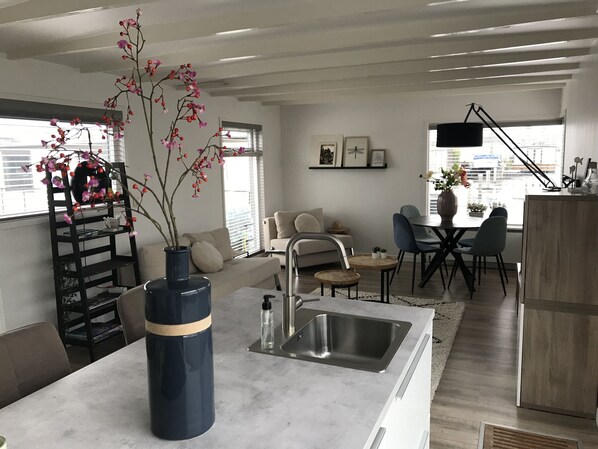 Una cocina moderna con un lavamanos integrado, una zona de comedor con mesa y sillas, un sofá y una planta decorativa en un jarrón.