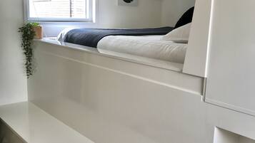 Une chambre à coucher comprenant un lit, une fenêtre, une étagère fixée au mur avec une plante, et deux éléments de décoration murale.