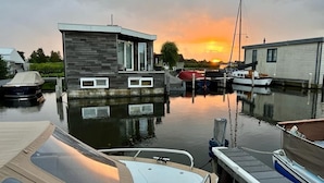 Una moderna casa flotante amarrada en un puerto deportivo con un atardecer de fondo.