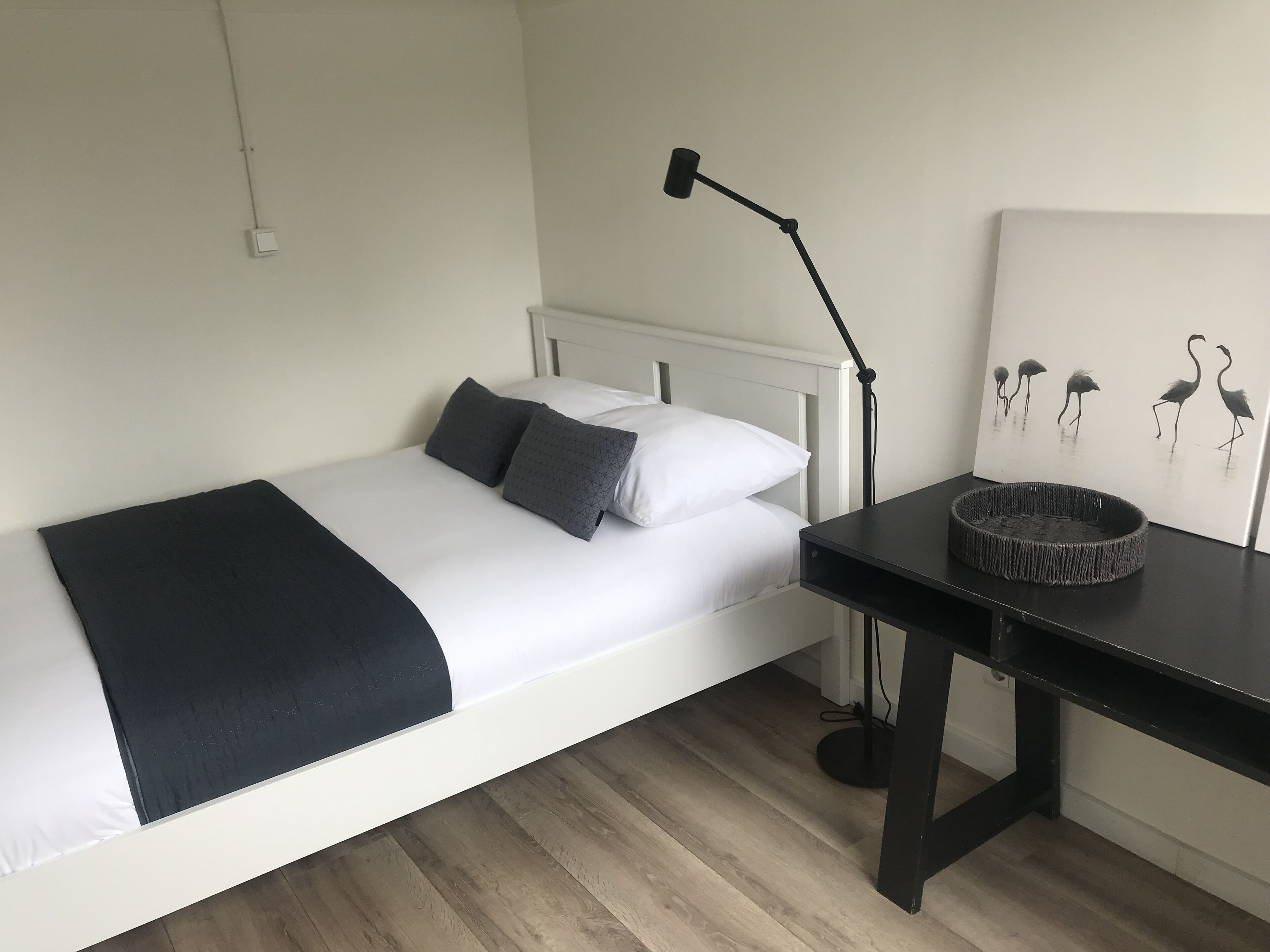 A neatly made bed with white and dark gray bedding, a black desk with a woven basket, and a wall-mounted lamp.