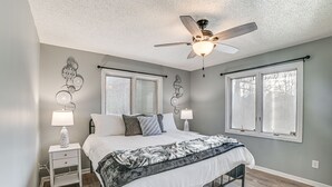 Une chambre à coucher comprenant un lit, deux tables de chevet, un ventilateur de plafond et deux fenêtres équipées de stores.