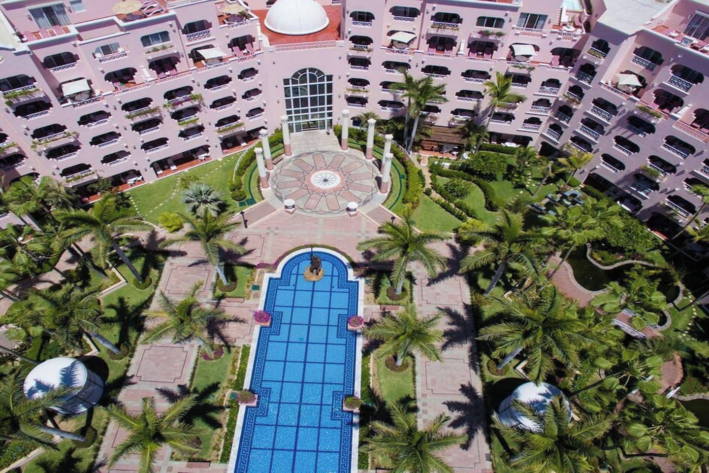 A multi-story hotel with a central courtyard featuring a swimming pool, surrounded by palm trees and a circular fountain.